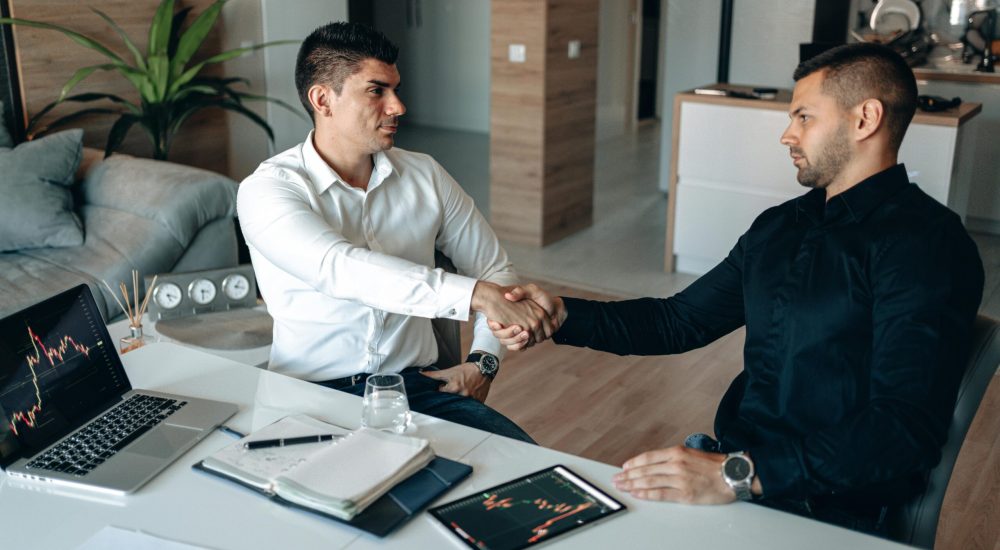 Two businessmen shake hands over financial charts and gadgets, symbolizing a successful deal.